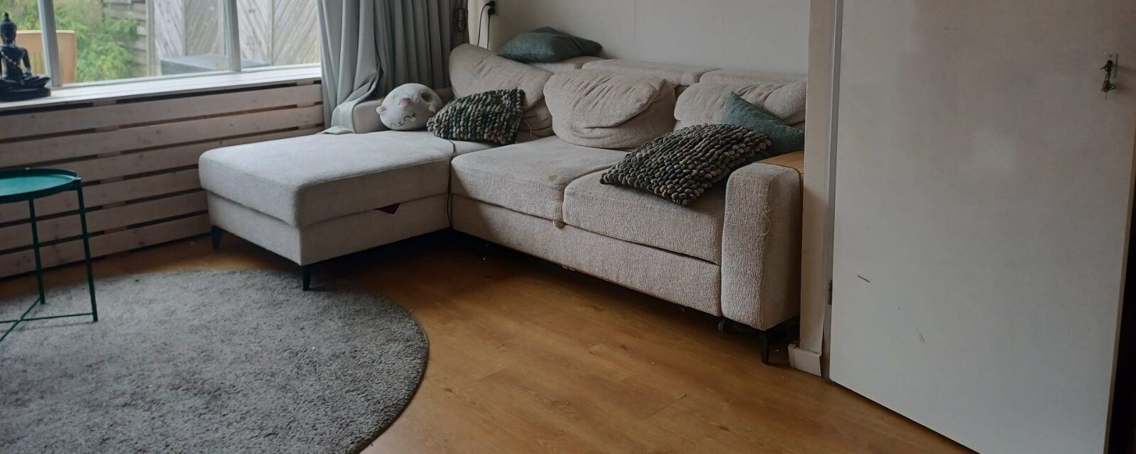 Beige hoekbank in een woonkamer, van een afstand gefotografeerd met de kussens erop, waardoor de vervuiling niet zichtbaar is